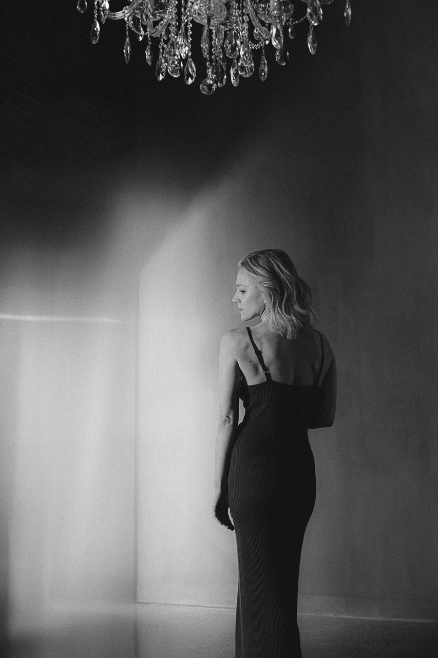 A woman with her back to the camera in a long fitted black dress, standing in a softly lit room beneath a crystal chandelier