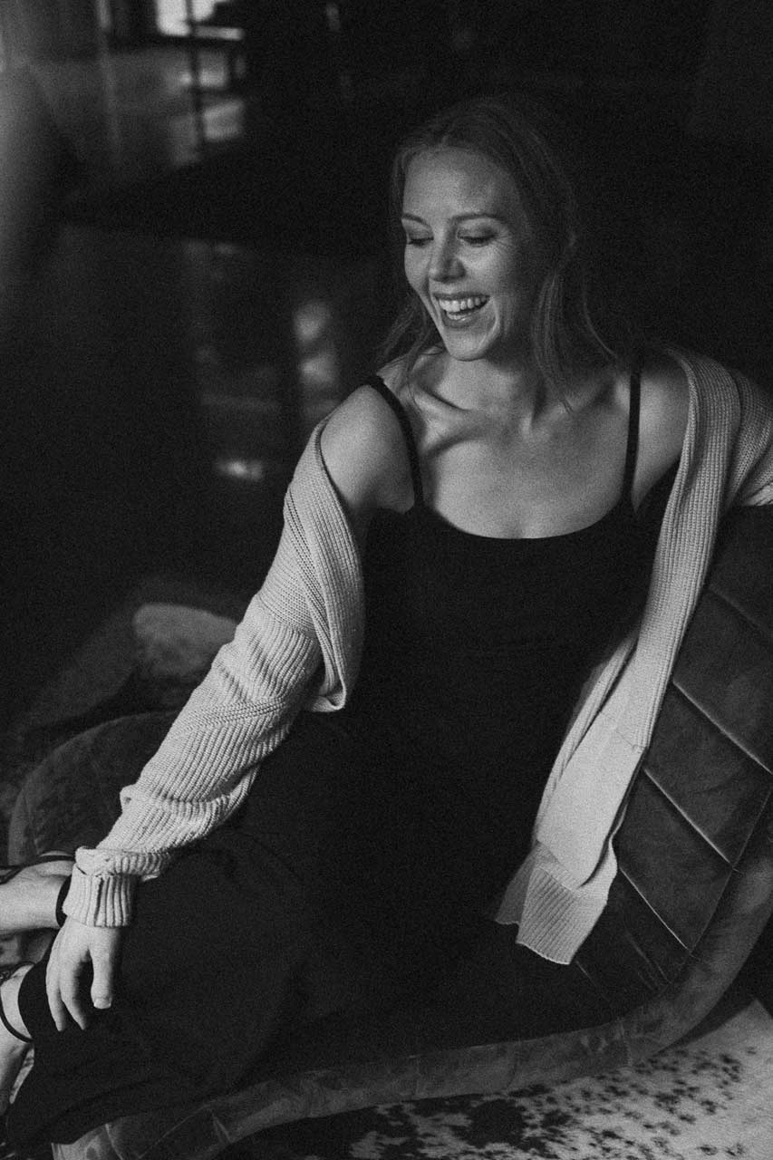 A woman laughing, curled up in a leather armchair with a knit cardigan slipping off her shoulder