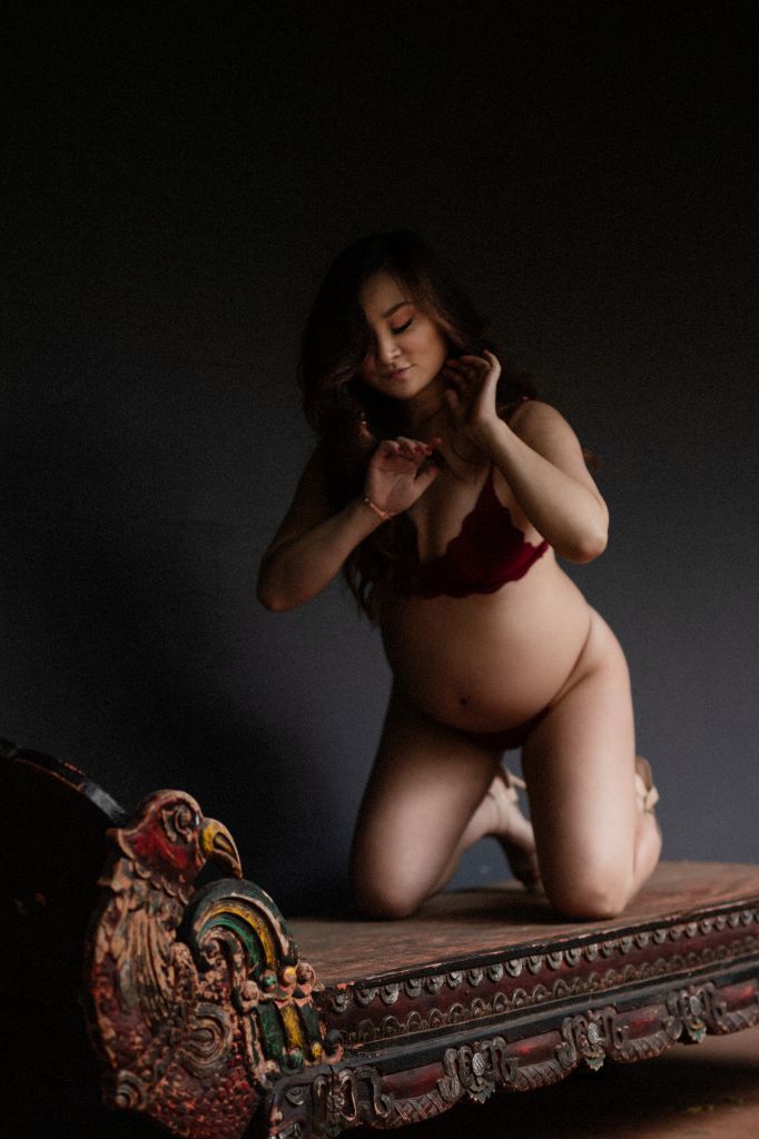 Expectant mother kneeling on a carved daybed in the studio, her bump lit against a dark backdrop