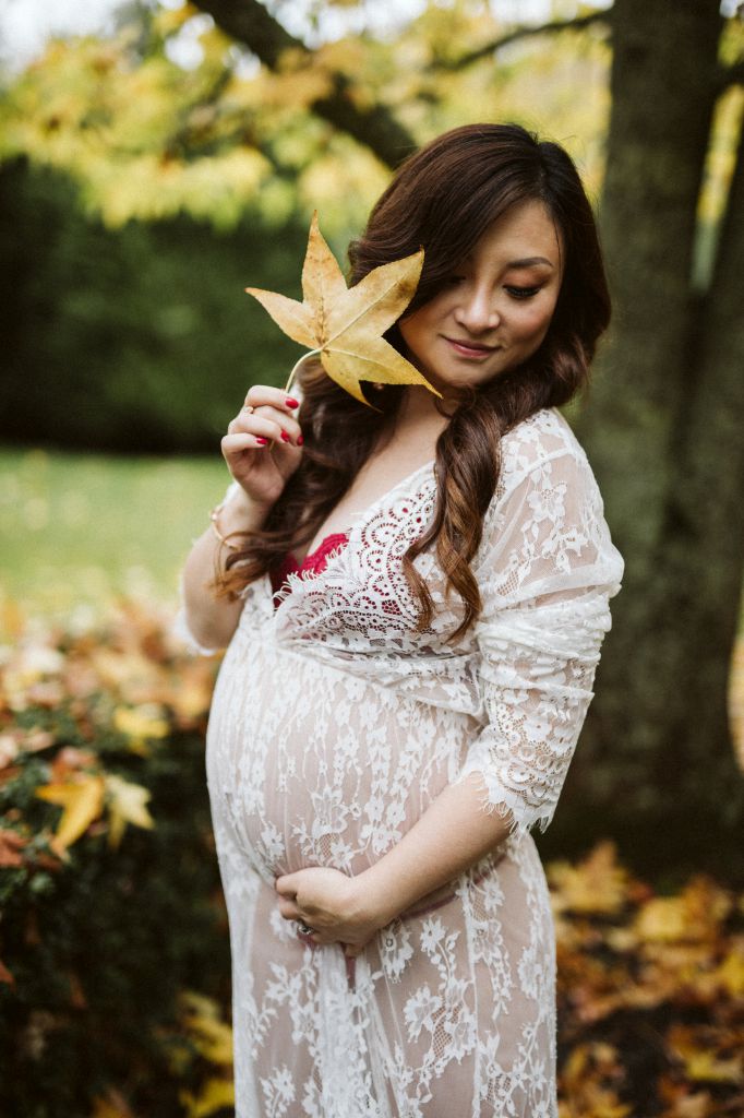 Expectant mother in a white lace gown cradling her bump, holding an autumn leaf beside her face